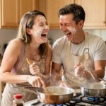 Paar lacht zusammen beim Kochen – Alltagsroutine als Beziehungskiller bekämpfen